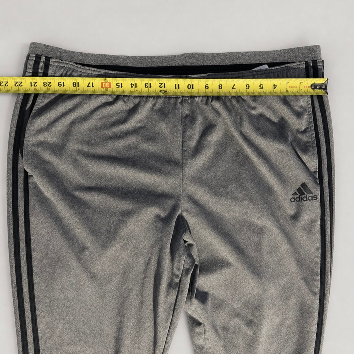 Gray Adidas track pants with black side stripes, shown with a yellow measuring tape across the waistband to indicate size.