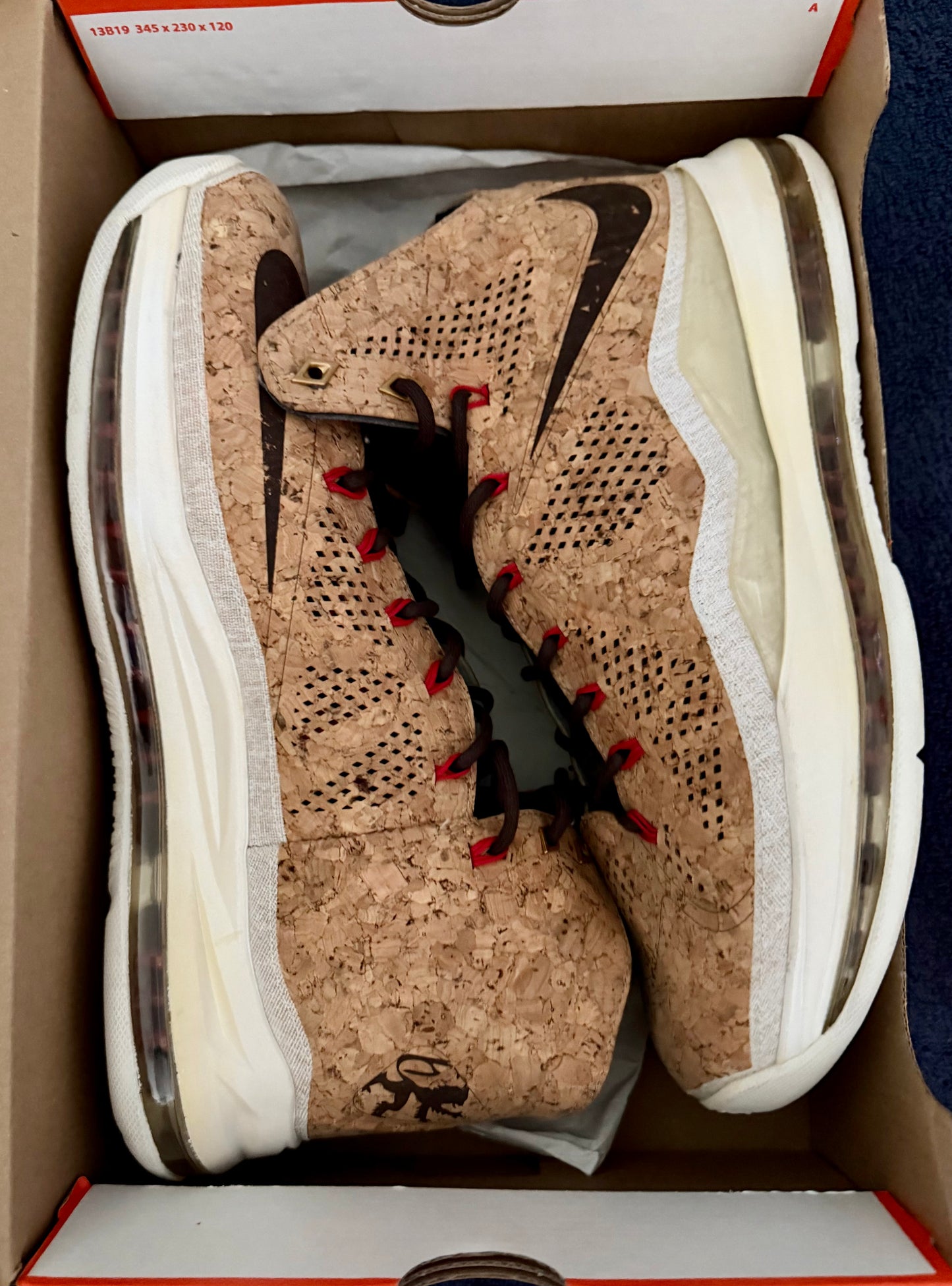 A pair of high-top sneakers with a cork-textured upper, black swooshes, and red laces, displayed inside their original white and red Nike box.
