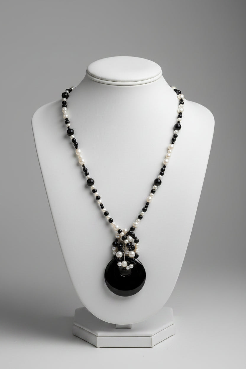 A beaded necklace with alternating black and white beads, featuring a dark oval pendant with a small silver-toned charm, displayed on a textured white fabric background.