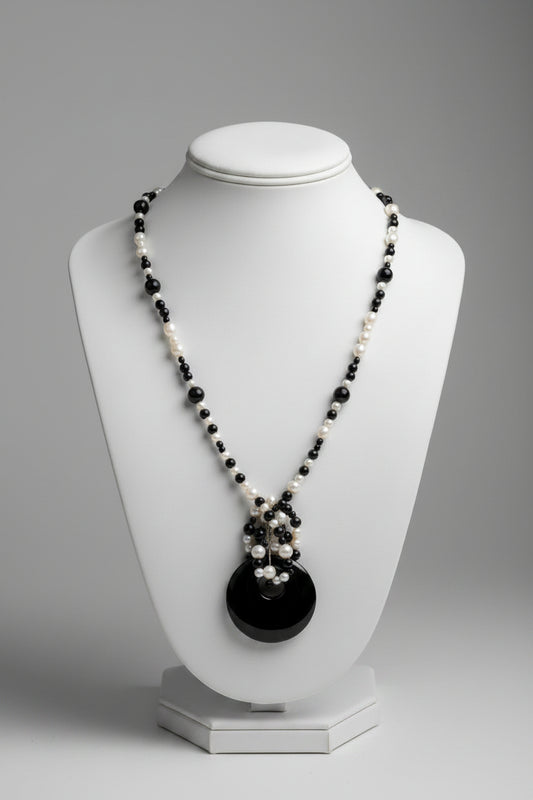 A beaded necklace with alternating black and white beads, featuring a dark oval pendant with a small silver-toned charm, displayed on a textured white fabric background.