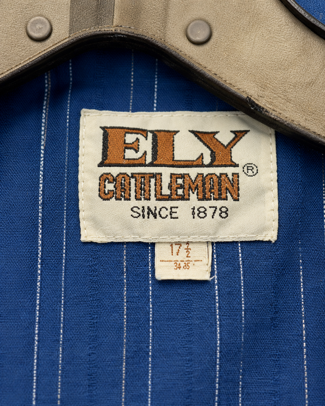 Close-up of a blue pinstriped shirt with a tan collar, featuring a visible embroidered brand label that reads 'ELY CATTLEMAN SINCE 1878'.