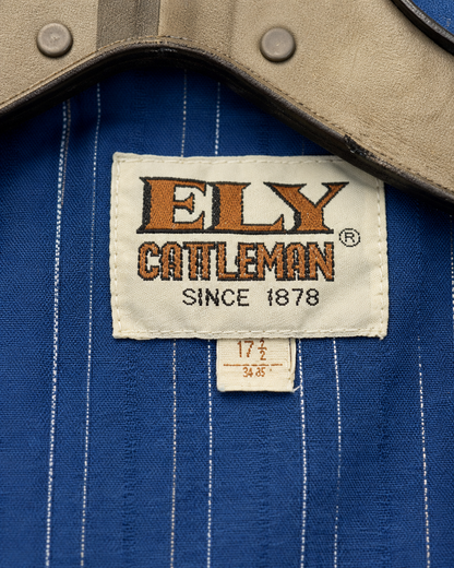 Close-up of a blue pinstriped shirt with a tan collar, featuring a visible embroidered brand label that reads 'ELY CATTLEMAN SINCE 1878'.