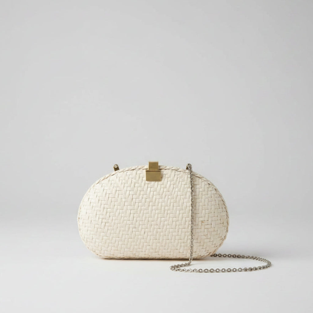 A small oval woven clutch bag in off-white with a gold-tone clasp and a silver-tone chain strap, hanging over a patterned rug in an indoor setting.