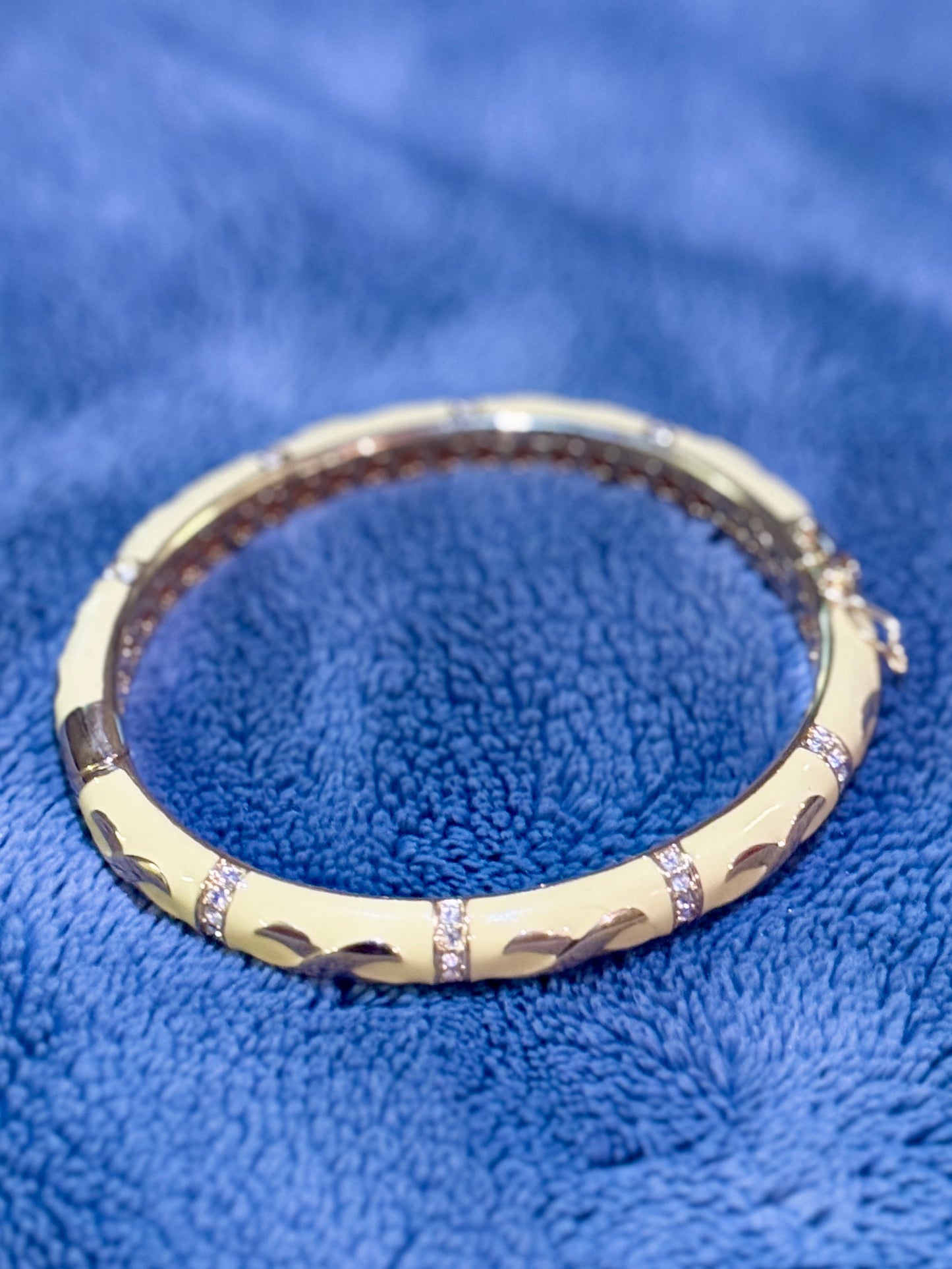 A gold bangle bracelet with a textured, wavy pattern and small clear crystal accents, displayed on a textured blue fabric background.
