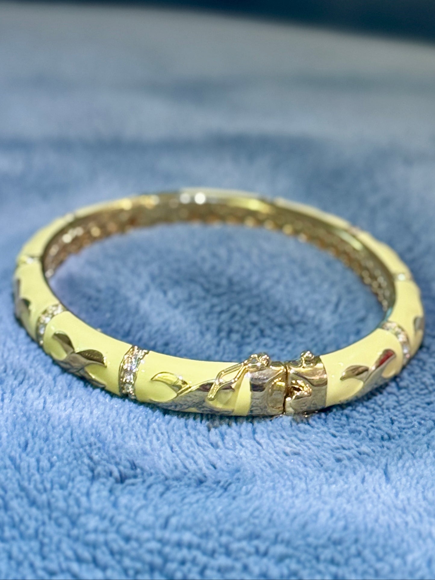 A gold-plated bangle bracelet with a textured leaf pattern and small clear rhinestone accents, resting on a soft blue textured fabric background.