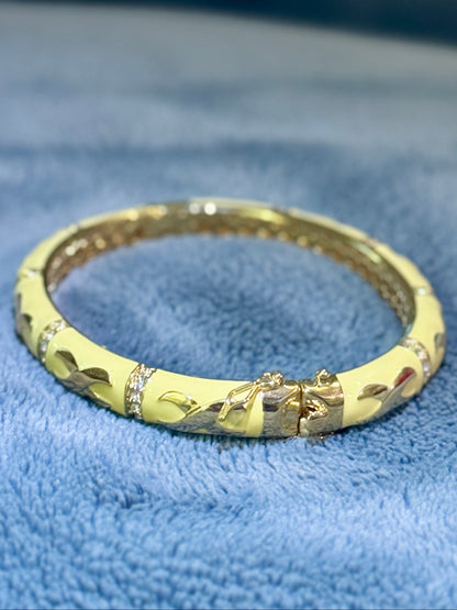 A gold-plated bangle bracelet with a textured leaf pattern and small clear rhinestone accents, resting on a soft blue textured fabric background.