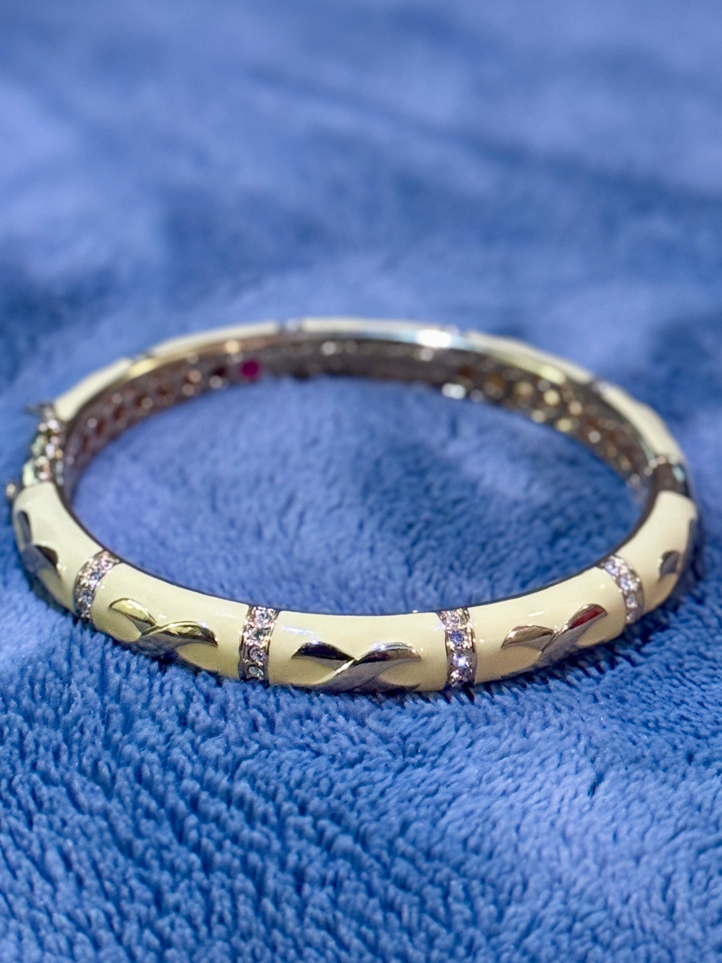 A gold-plated bangle bracelet with a cream-colored central inlay featuring stylized leaf or wing motifs, accented by rows of small clear stones, resting on a textured blue fabric background.