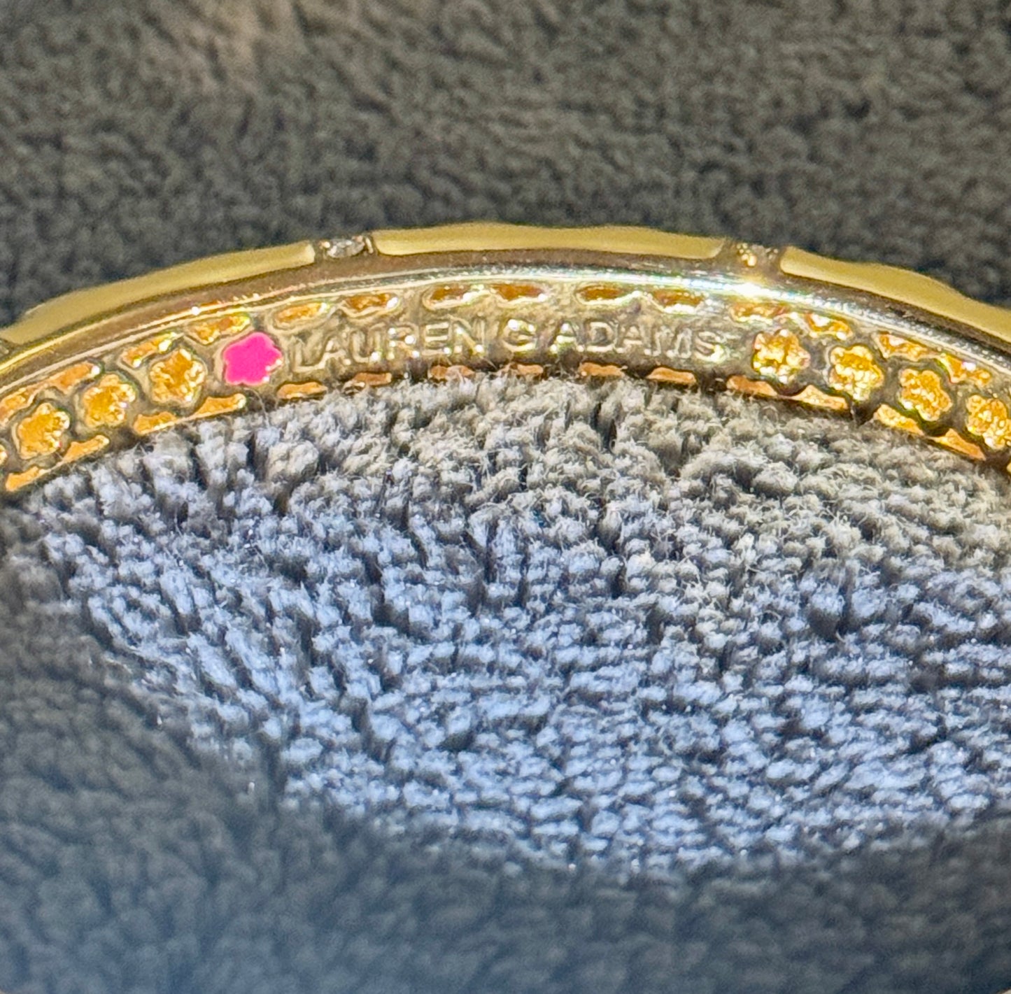Close-up of a gold bangle bracelet with floral cutout detailing, a small pink gemstone, and 'LAUREN ADAMS' engraved on the inside, resting on a textured gray fabric surface.
