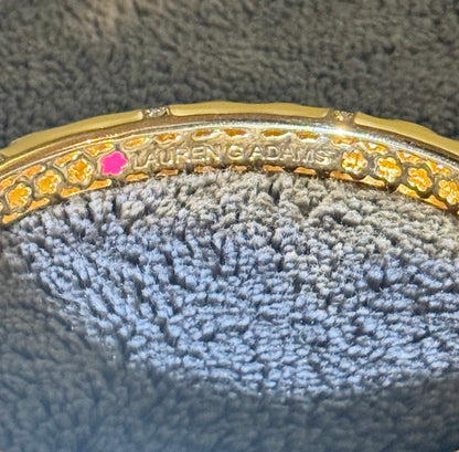 Close-up of a gold bangle bracelet with floral cutout detailing, a small pink gemstone, and 'LAUREN ADAMS' engraved on the inside, resting on a textured gray fabric surface.