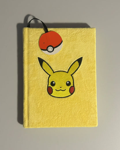 A yellow notebook or journal featuring a printed Pikachu face and a red-and-white Pokéball bookmark attached near the top, placed on a textured gray fabric surface.