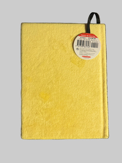 A yellow hardcover notebook with a textured cover and a black ribbon bookmark, resting on a gray textured blanket. A circular product tag is attached near the top right.