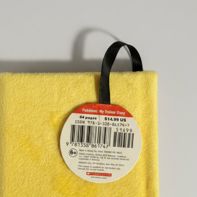 A close-up of a bright yellow Pokémon TCG card deck box with a black hanging loop and a round white price tag reading 'Pokémon: My Trainer Story' and '64 pages $14.99 US'.