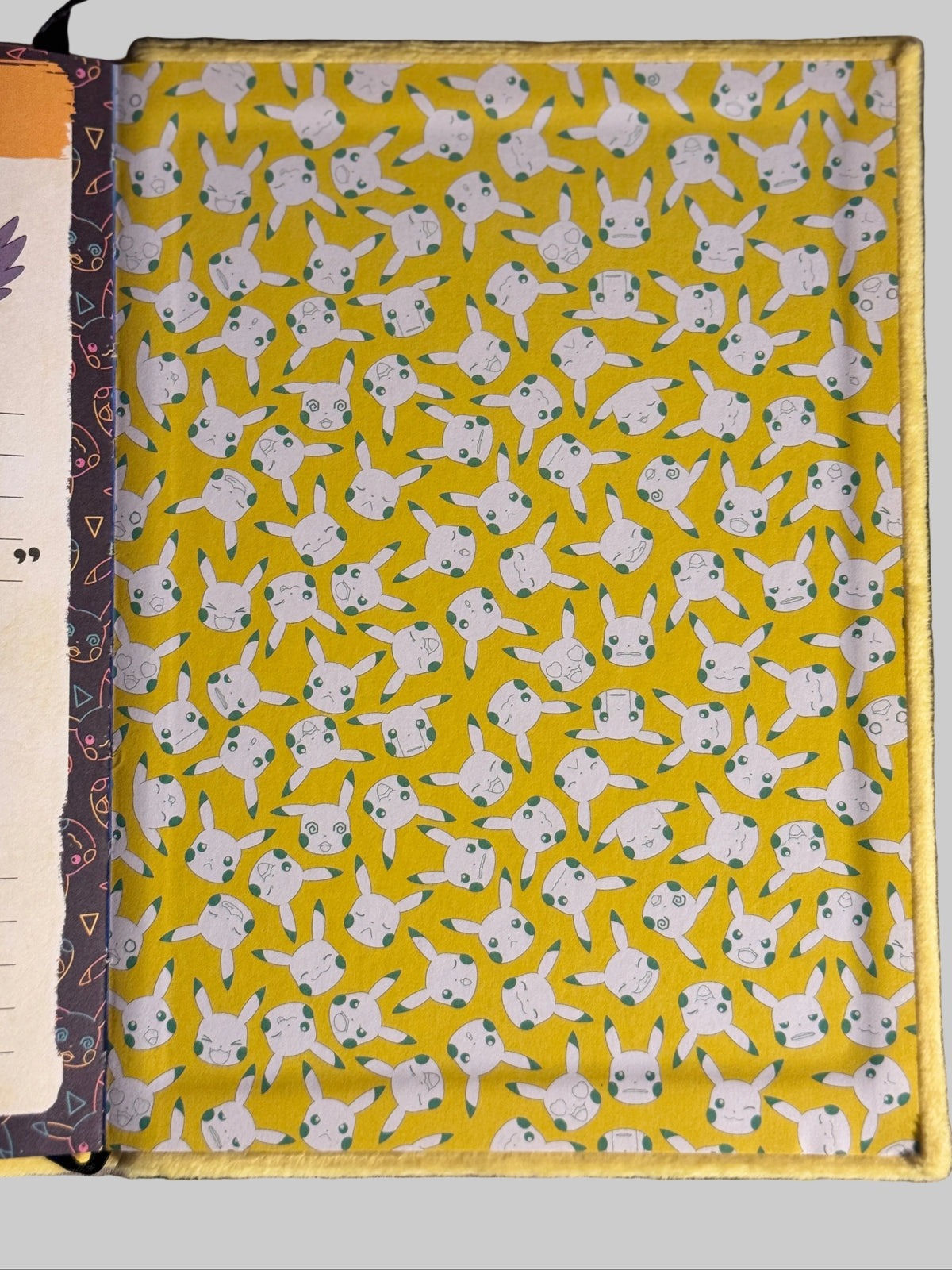 A close-up of a fabric-covered notebook or journal cover laid flat on a gray textured surface. The visible cover has a bright yellow background with an allover repeating print of white cartoon characters with small blue accents.
