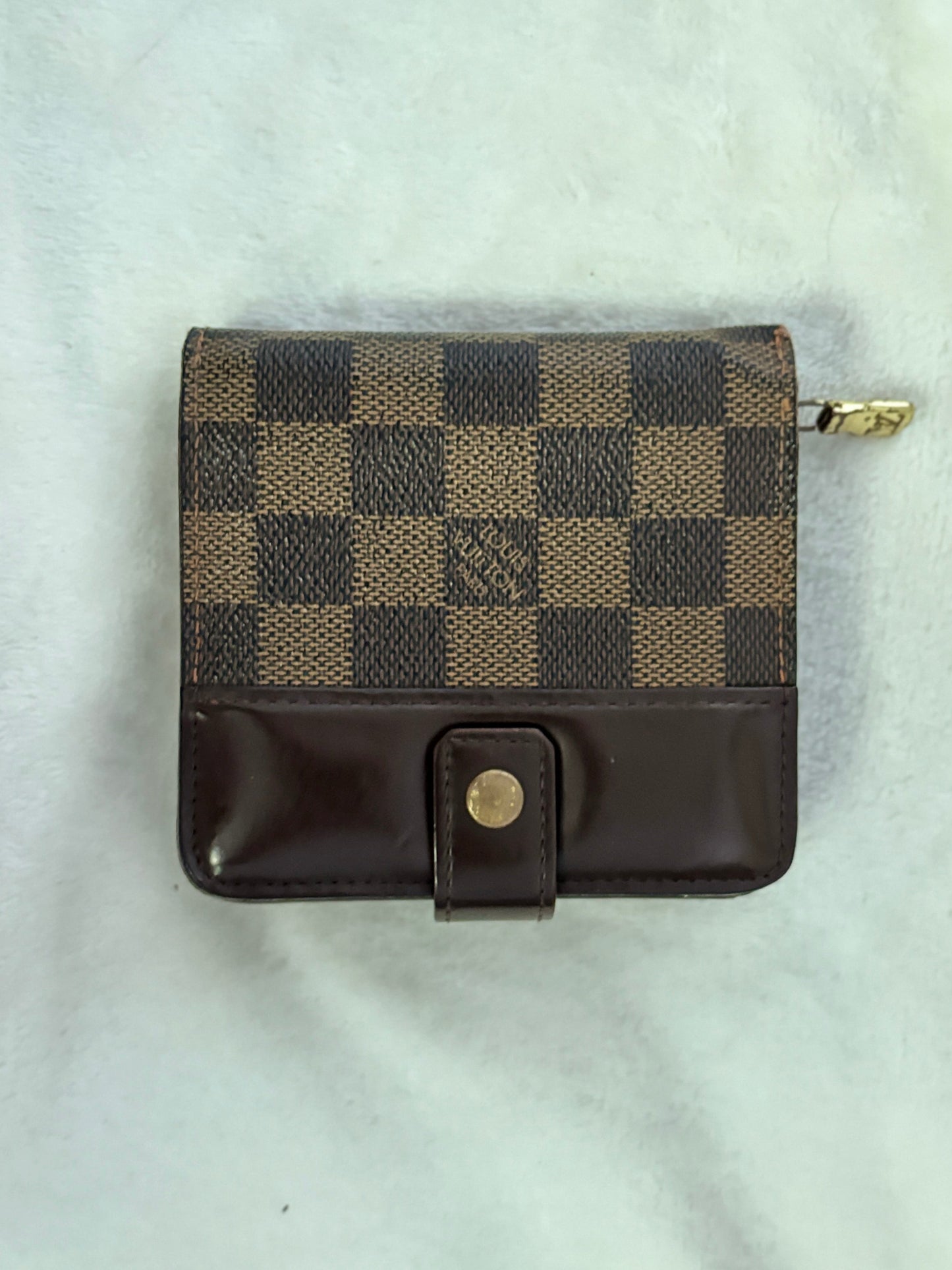 A small Louis Vuitton zip-around wallet in the Damier Ebene checkered canvas and dark brown leather, featuring a snap-button closure, laid flat on a white textured fabric background.