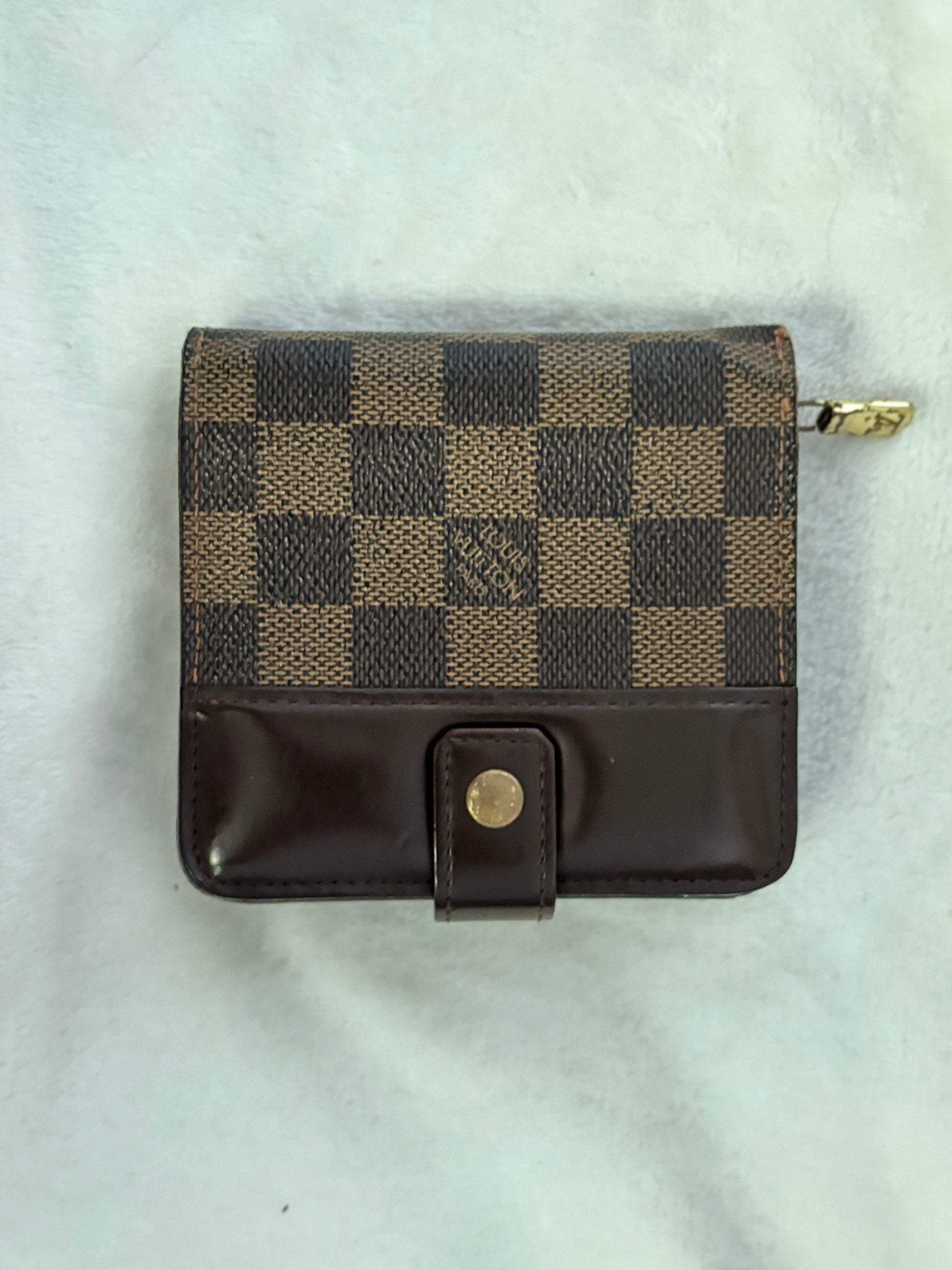 A small Louis Vuitton zip-around wallet in the Damier Ebene checkered canvas and dark brown leather, featuring a snap-button closure, laid flat on a white textured fabric background.