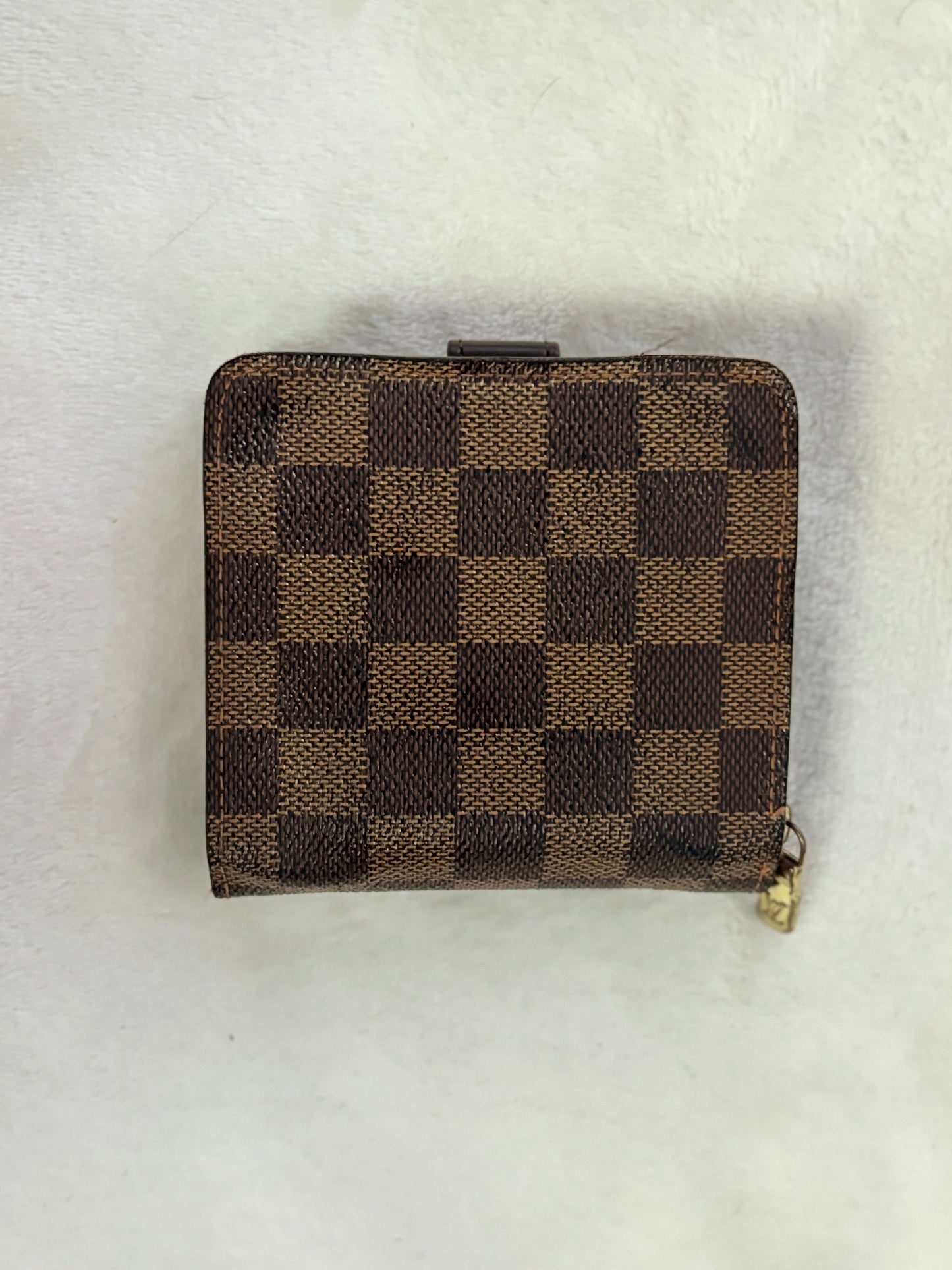 A small brown and tan checkered wallet with a gold zipper pull, placed on a white textured background.