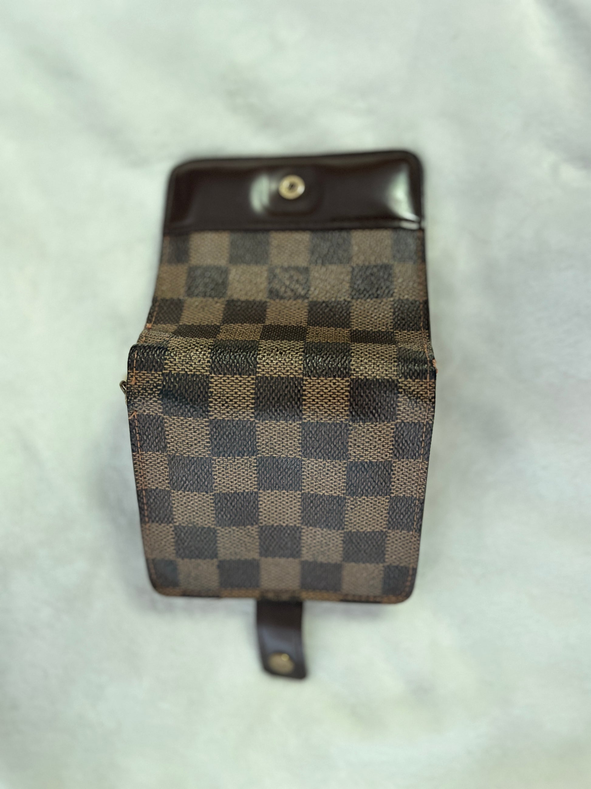 A small checkered pouch or wallet with a dark brown leather top and strap, featuring a brown and beige checkerboard pattern, photographed on a light textured fabric background.