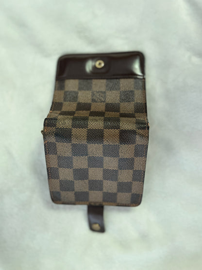 A small checkered pouch or wallet with a dark brown leather top and strap, featuring a brown and beige checkerboard pattern, photographed on a light textured fabric background.