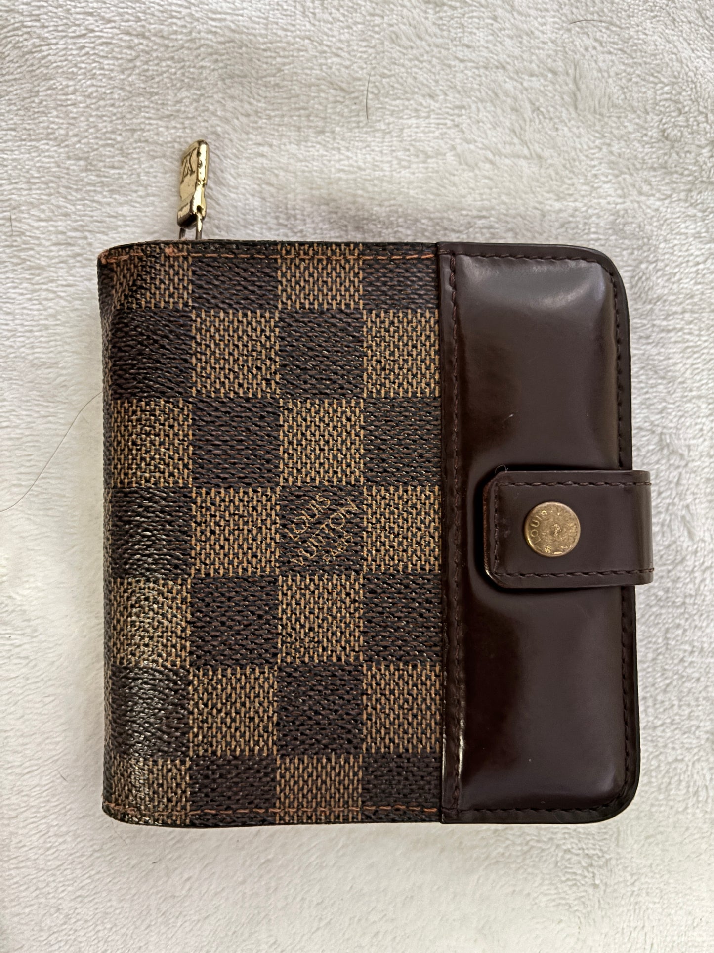 A Louis Vuitton zip-around wallet in the Damier Ebene checkered canvas with a dark brown leather flap and snap button closure, resting on a white textured fabric background.