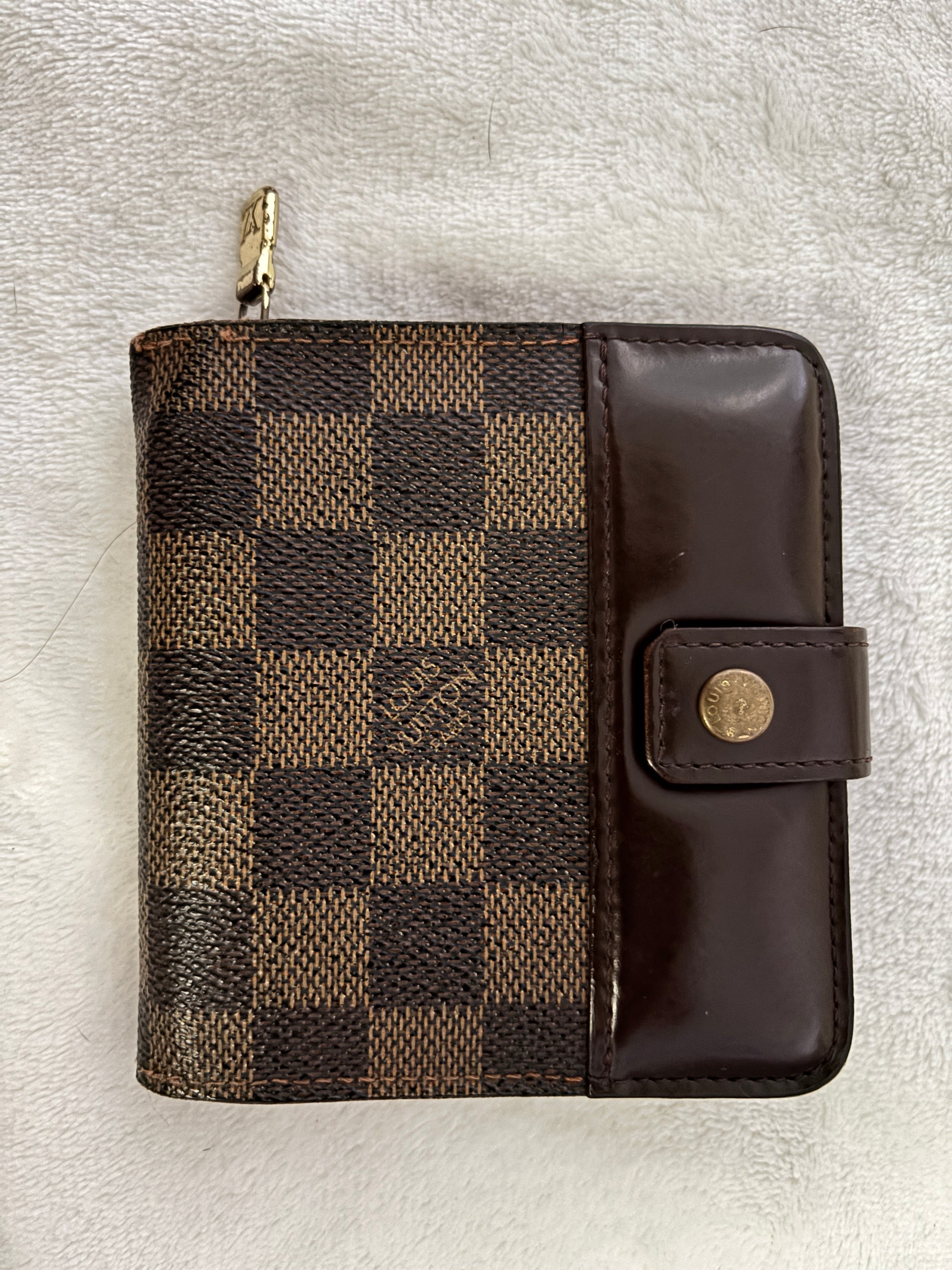 A Louis Vuitton zip-around wallet in the Damier Ebene checkered canvas with a dark brown leather flap and snap button closure, resting on a white textured fabric background.