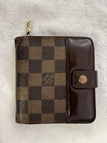 A Louis Vuitton zip-around wallet in the Damier Ebene checkered canvas with a dark brown leather flap and snap button closure, resting on a white textured fabric background.