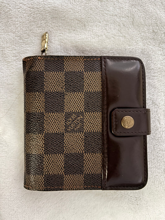 A Louis Vuitton zip-around wallet in the Damier Ebene checkered canvas with a dark brown leather flap and snap button closure, resting on a white textured fabric background.