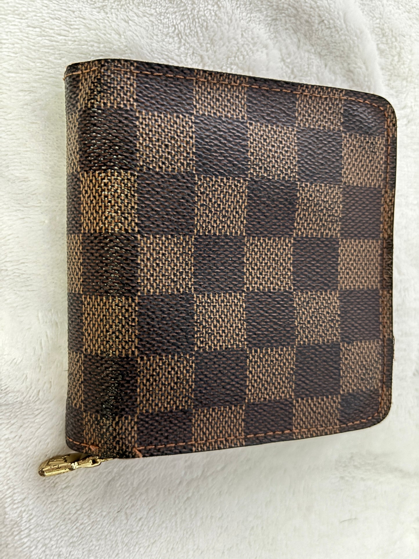 A Louis Vuitton zip-around wallet in the Damier Ebene checkered canvas, featuring a brown and beige checkerboard pattern with a gold-tone zipper pull, resting on a white textured fabric background.