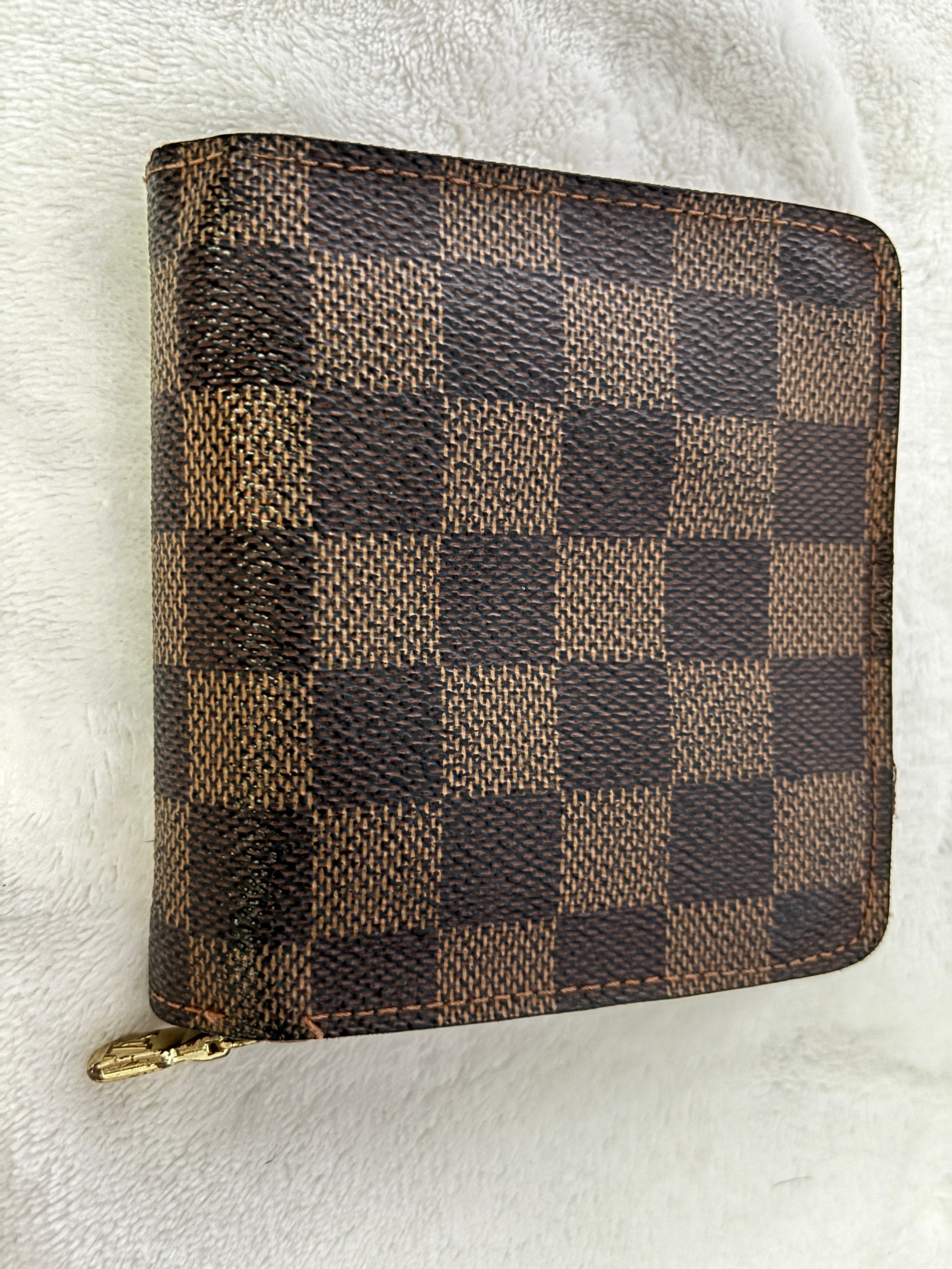 A Louis Vuitton zip-around wallet in the Damier Ebene checkered canvas, featuring a brown and beige checkerboard pattern with a gold-tone zipper pull, resting on a white textured fabric background.
