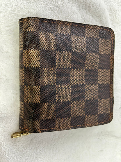 A Louis Vuitton zip-around wallet in the Damier Ebene checkered canvas, featuring a brown and beige checkerboard pattern with a gold-tone zipper pull, resting on a white textured fabric background.