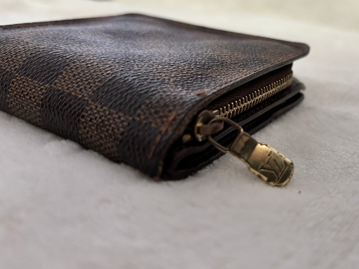 A close-up shot of a brown textured leather wallet with a gold-tone zipper pull, resting on a soft, light-colored fabric surface.