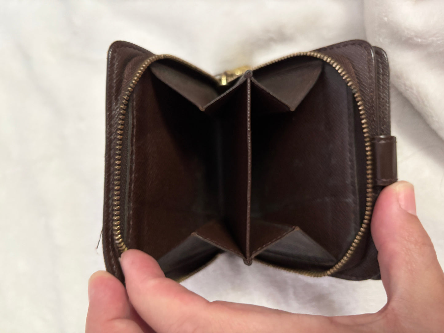 An open dark brown leather wallet held in two hands, showing its interior compartments and gold-toned zipper against a white textured fabric background.