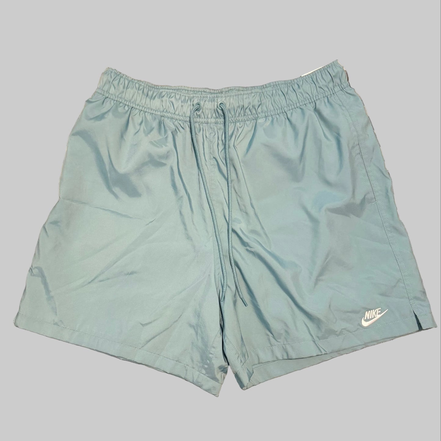 Light blue Nike athletic shorts laid flat on a gray and white plaid bedsheet, with a person's legs and white shirt visible at the bottom of the frame.