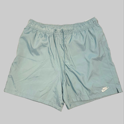 Light blue Nike athletic shorts laid flat on a gray and white plaid bedsheet, with a person's legs and white shirt visible at the bottom of the frame.