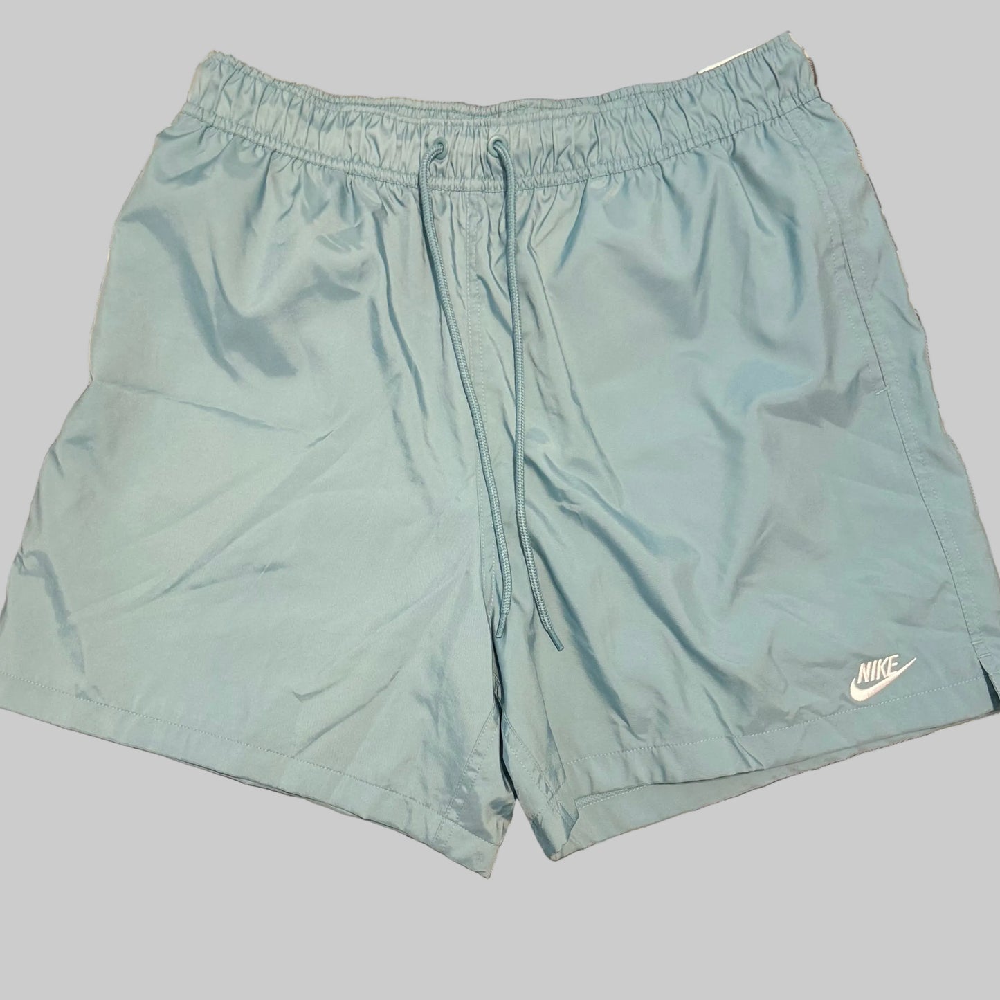 Light blue Nike athletic shorts laid flat on a gray and white plaid bedsheet, with a person's legs and white shirt visible at the bottom of the frame.