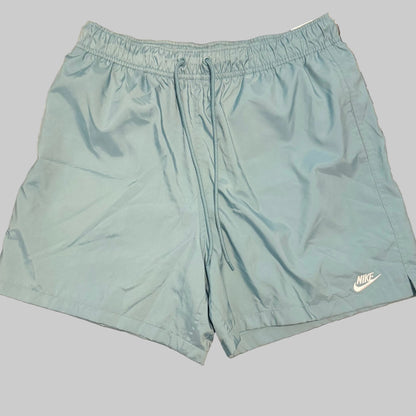 Light blue Nike athletic shorts laid flat on a gray and white plaid bedsheet, with a person's legs and white shirt visible at the bottom of the frame.