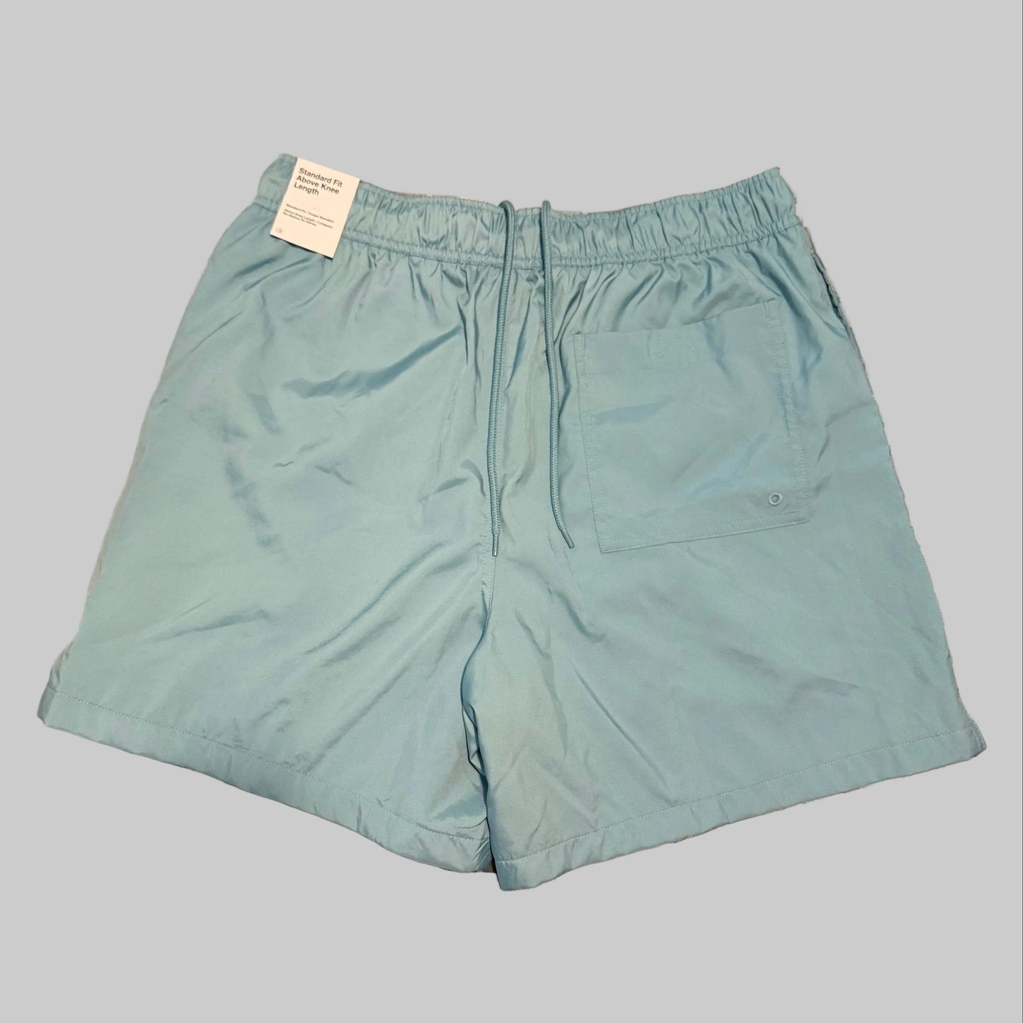 Light blue swim trunks laid flat on a plaid bedsheet, with a white tag still attached at the waistband.