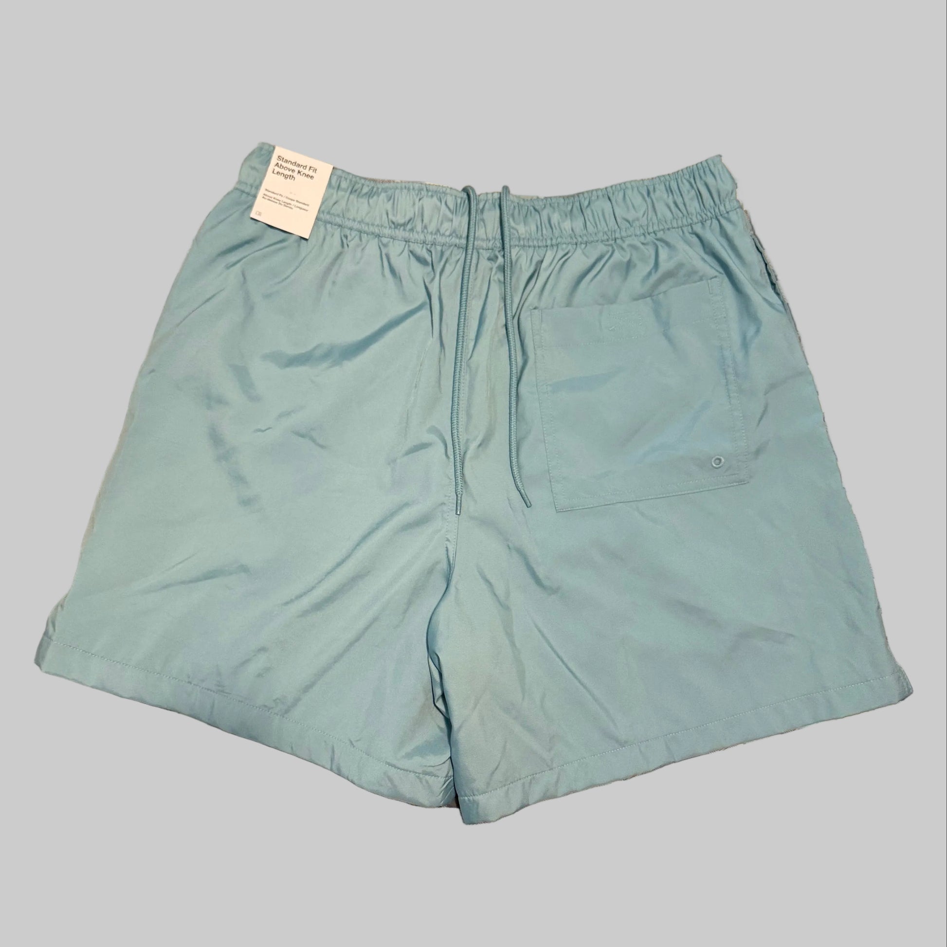 Light blue swim trunks laid flat on a plaid bedsheet, with a white tag still attached at the waistband.