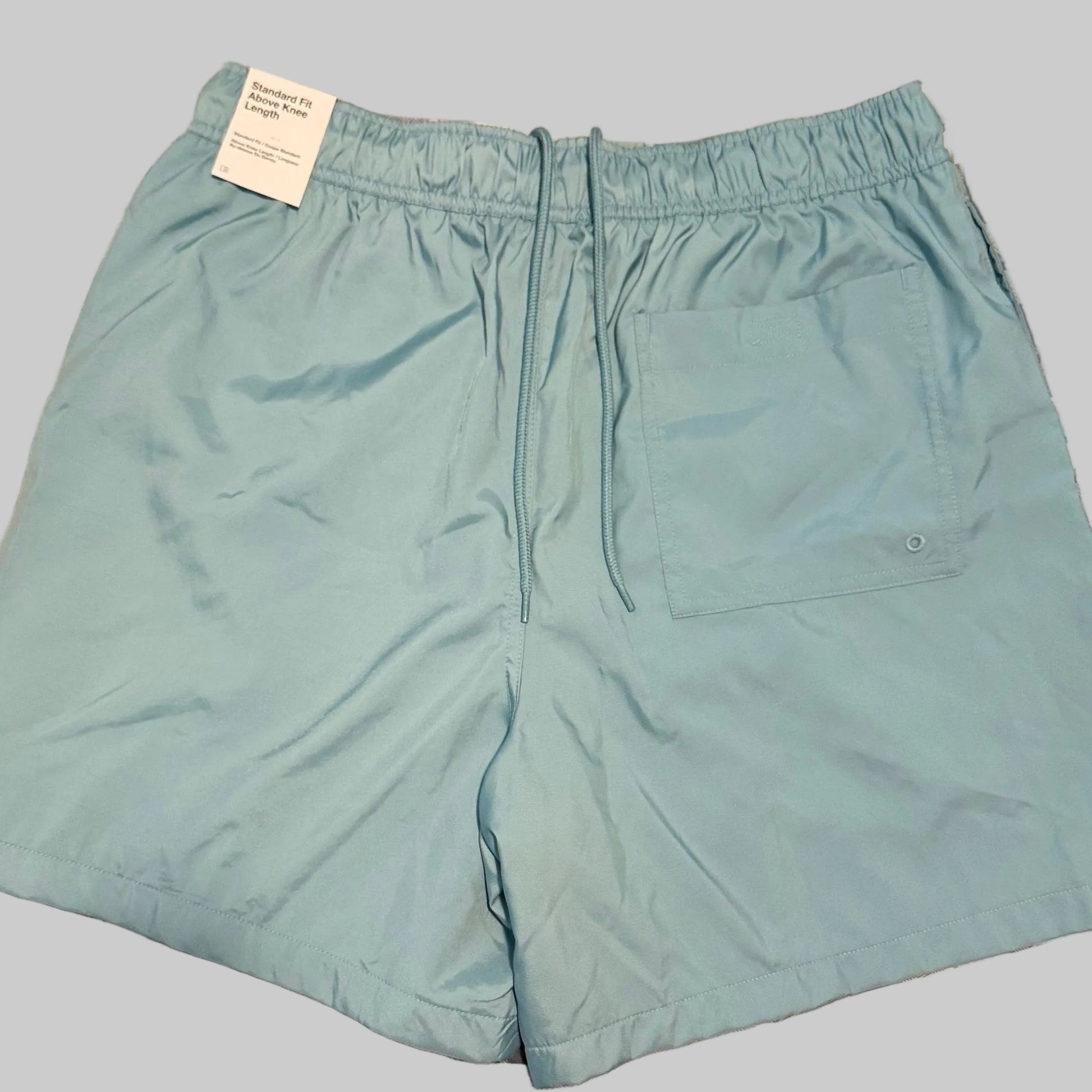Light blue swim trunks laid flat on a plaid bedsheet, with a white tag still attached at the waistband.