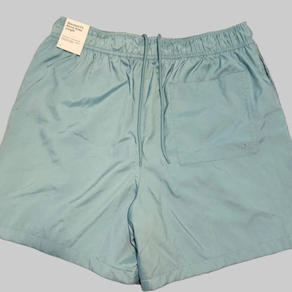 Light blue swim trunks laid flat on a plaid bedsheet, with a white tag still attached at the waistband.