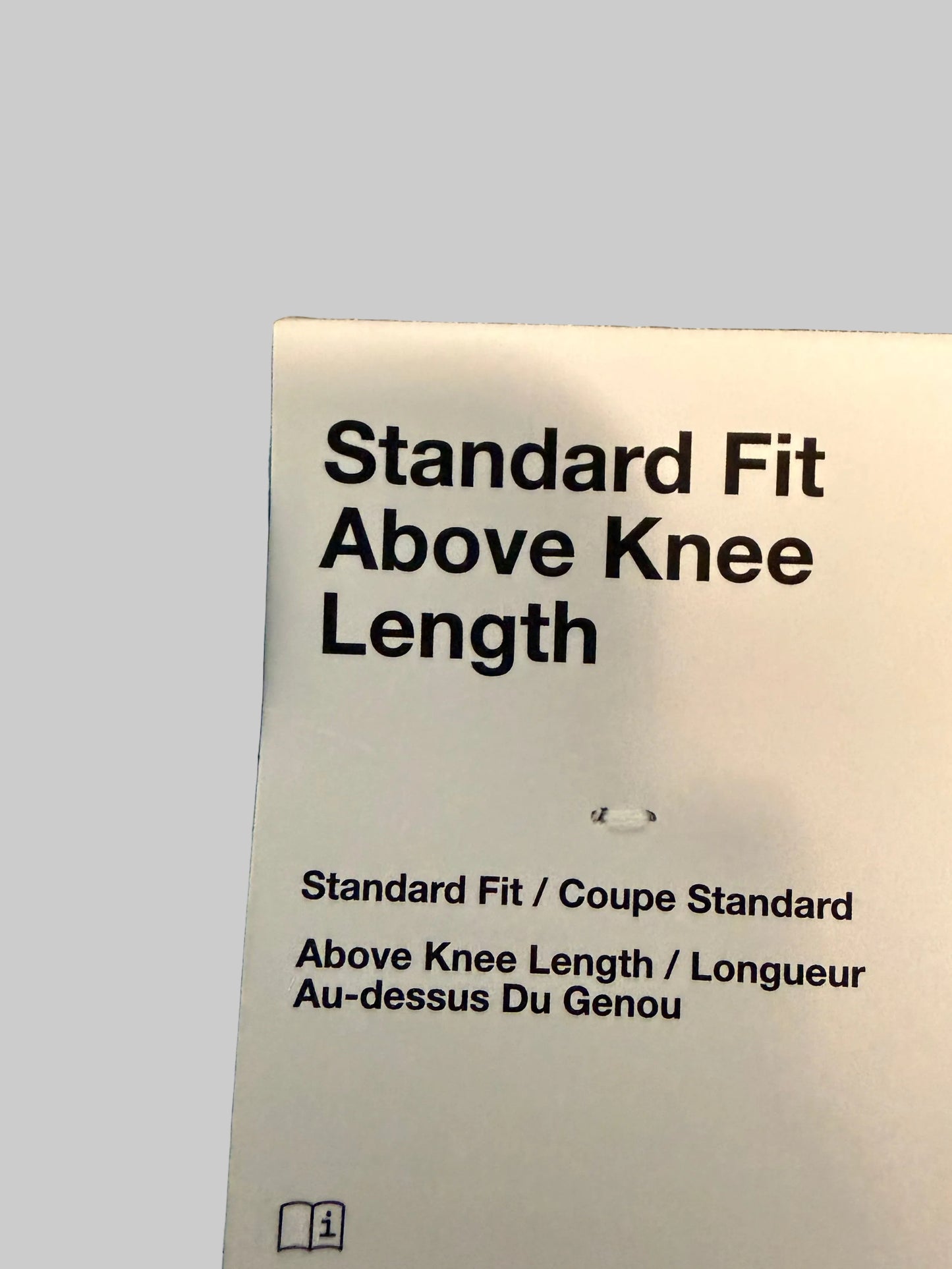 A white size label reading 'Standard Fit Above Knee Length' attached to light blue pants, photographed close up against a gray plaid fabric background.