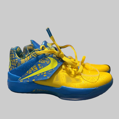 A pair of yellow and blue Nike basketball shoes resting on a gray and white plaid blanket in a bedroom setting.