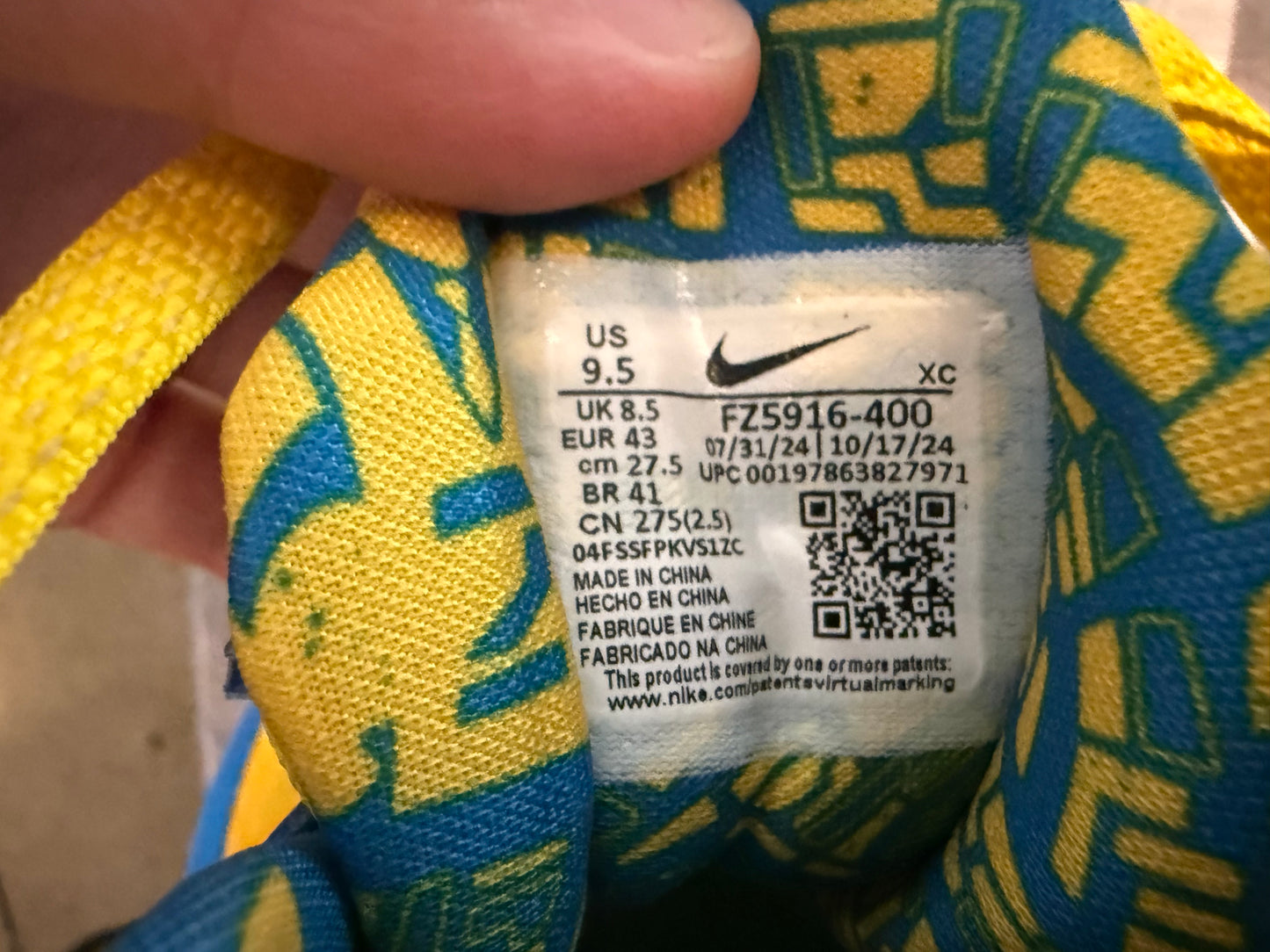 Close-up of a Nike shoe size tag on a blue and yellow patterned athletic shoe, with a finger holding the tag in place.