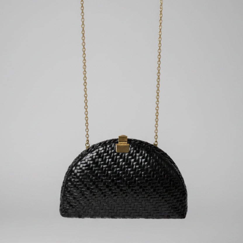 A small black woven handbag with a gold clasp and gold chain strap, photographed hanging in an indoor setting with a patterned rug and furniture visible in the background.
