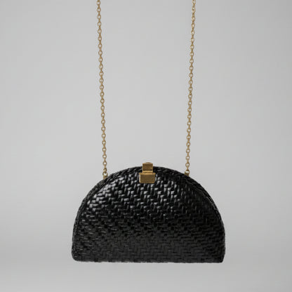 A small black woven handbag with a gold clasp and gold chain strap, photographed hanging in an indoor setting with a patterned rug and furniture visible in the background.