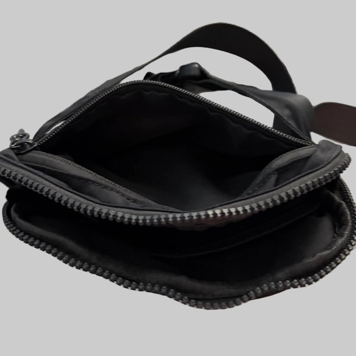 A black nylon crossbody bag shown open, displaying its interior lining and zipper compartments, set against a solid gray background.