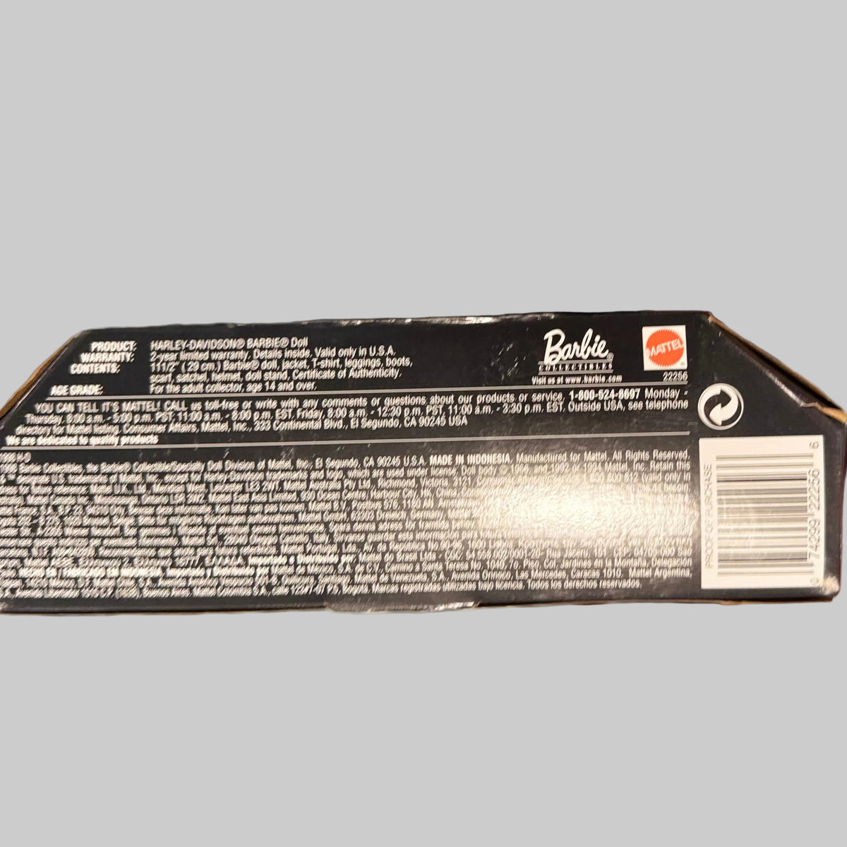 A hand holding a black Barbie-themed product box, showing the back with white text, a barcode, and a small orange logo, against a blurred blue-and-white striped background.