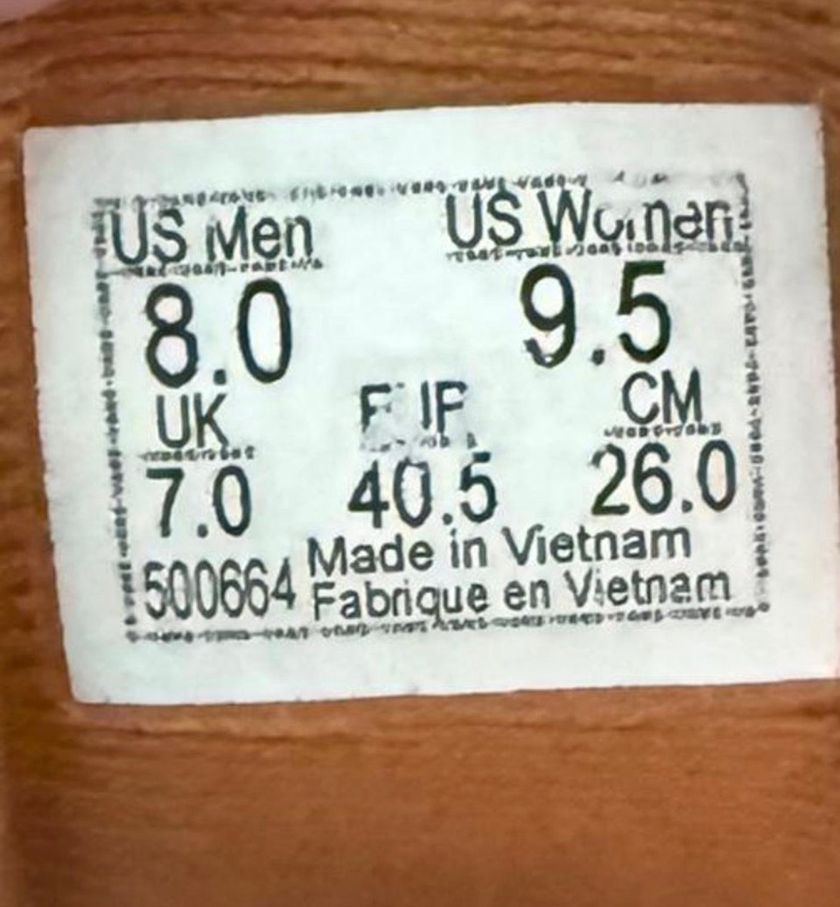 A white size chart sticker with black printed text showing US Men and US Women measurements in inches and centimeters, plus 'Made in Vietnam' and a product code, placed on a brown wooden surface.