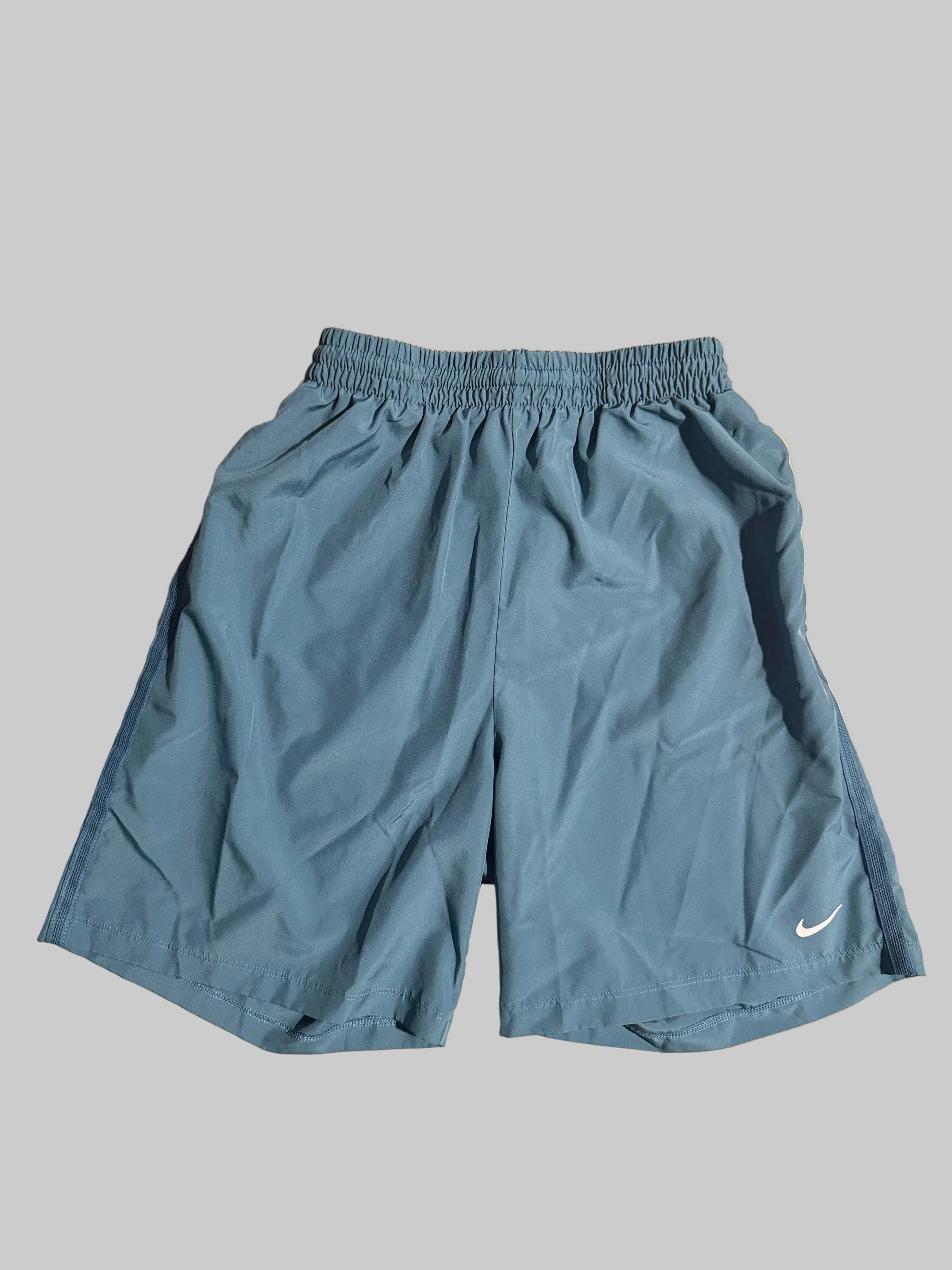 A pair of light blue athletic shorts with an elastic waistband and a small white Nike swoosh on the lower left leg, laid flat on a light-colored wooden surface.