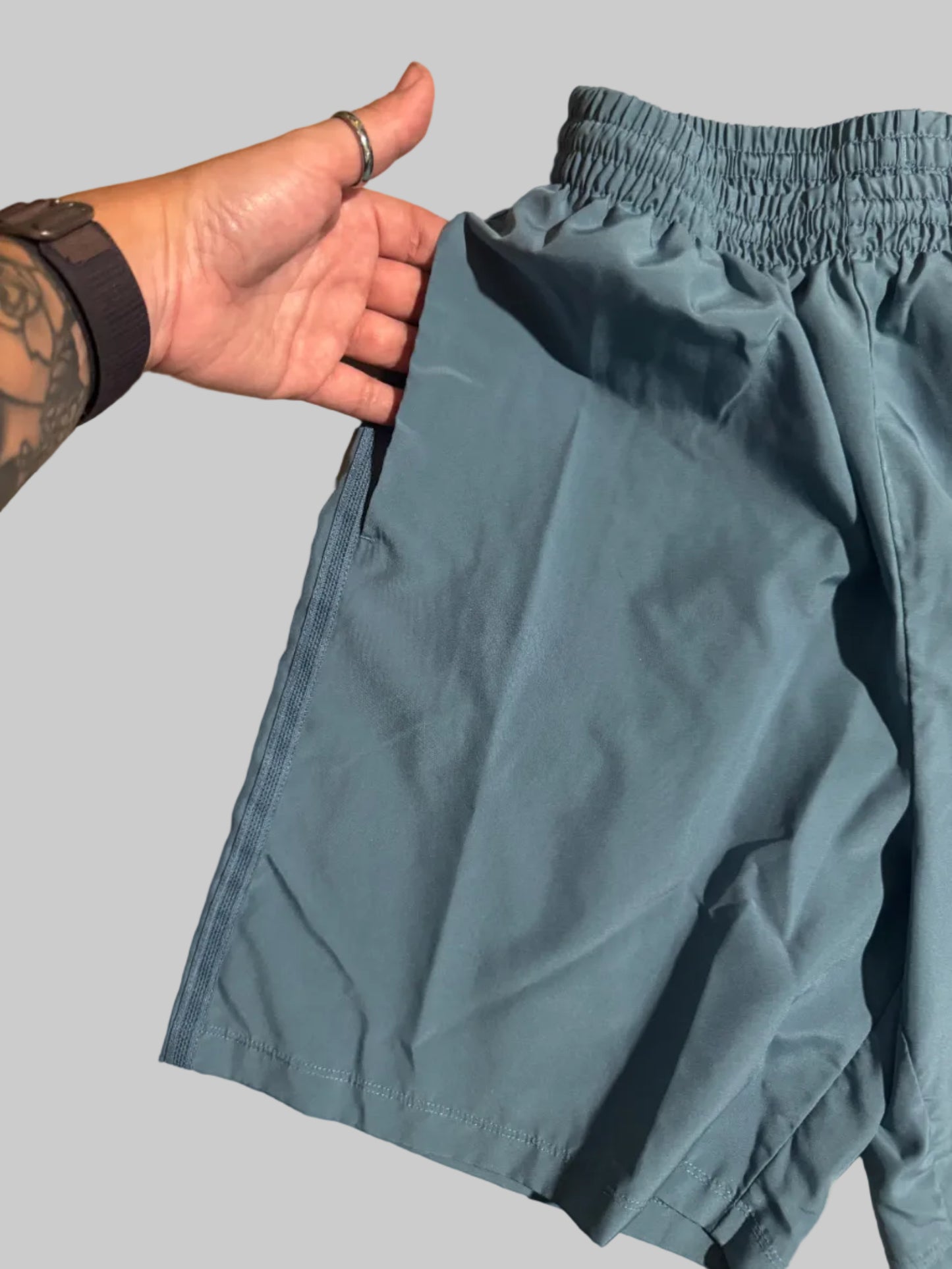 A pair of slate blue athletic shorts with an elastic waistband, shown with a person's hand holding the hem to demonstrate the fit on a beige tiled floor.