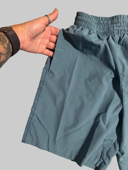 A pair of slate blue athletic shorts with an elastic waistband, shown with a person's hand holding the hem to demonstrate the fit on a beige tiled floor.